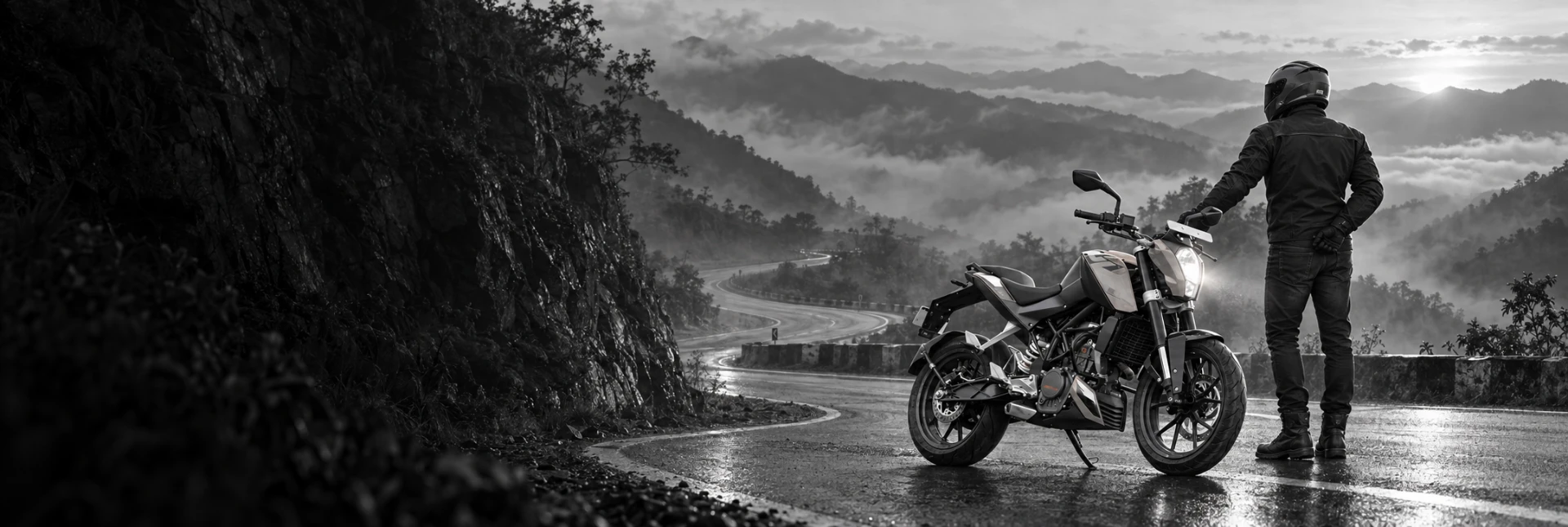 A helmeted motorcyclist standing beside a motorcycle on a wet mountain road overlooking misty hills